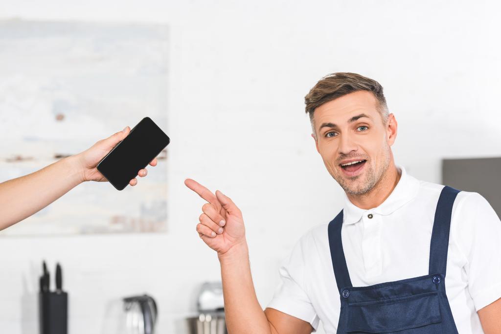 visão parcial da mão segurando smartphone e sorridente reparador adulto apontando para a tela em branco
  - Foto, Imagem