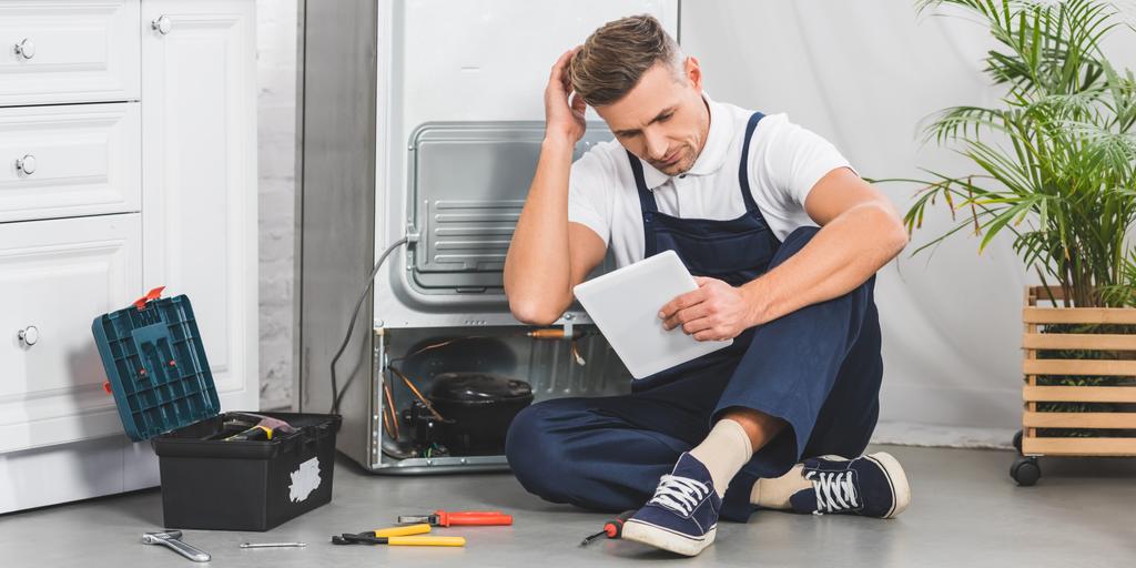 thoughtful adult repairman sitting on floor and using digital tablet while repairing refrigerator - Photo, Image