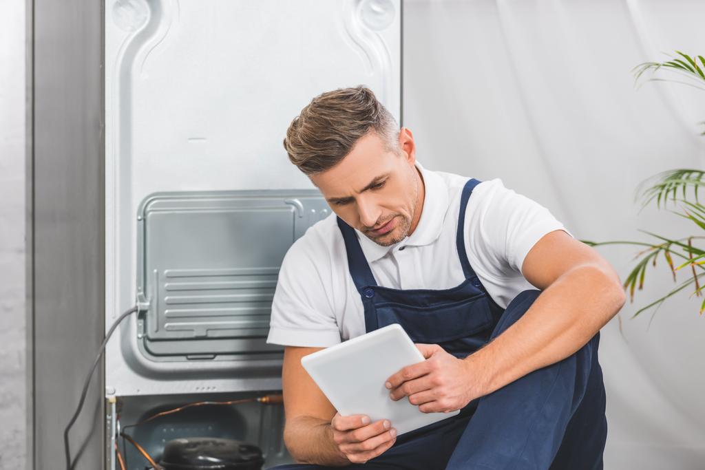 Erwachsener Reparateur sitzt auf dem Boden und benutzt digitales Tablet, während er Kühlschrank repariert - Foto, Bild