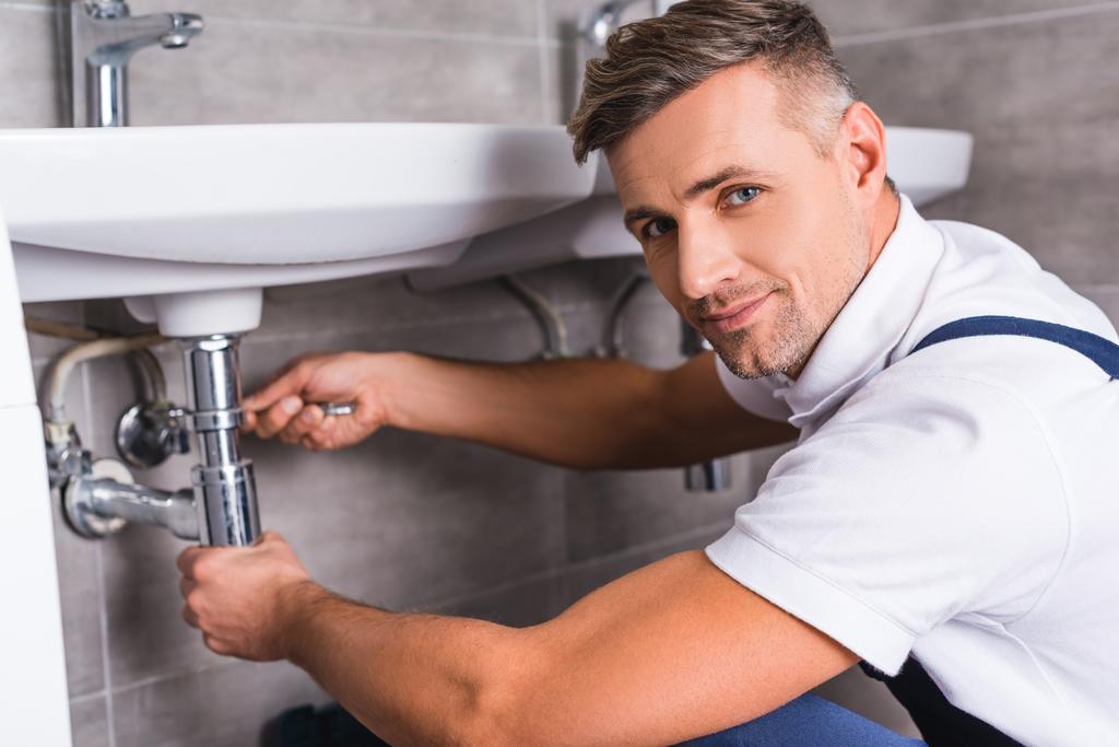adult repairman repairing sink at bathroom and looking at camera - Photo, Image