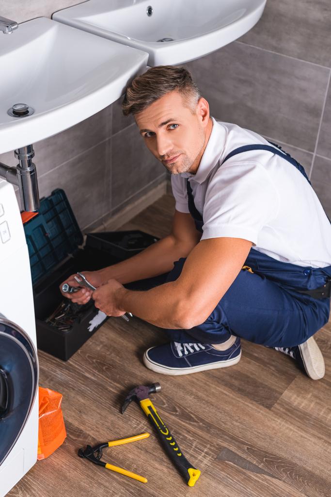 Hochwinkelaufnahme eines erwachsenen Reparateurs, der auf dem Boden sitzt, Reparaturwerkzeuge in der Hand hält und in die Kamera schaut - Foto, Bild