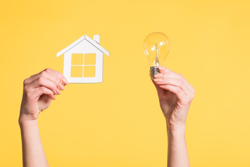 bijgesneden beeld van vrouwelijke handen houden papier huis en onder leiding van lamp in handen geïsoleerd op geel, energie-efficiëntie thuis concept - Foto, afbeelding