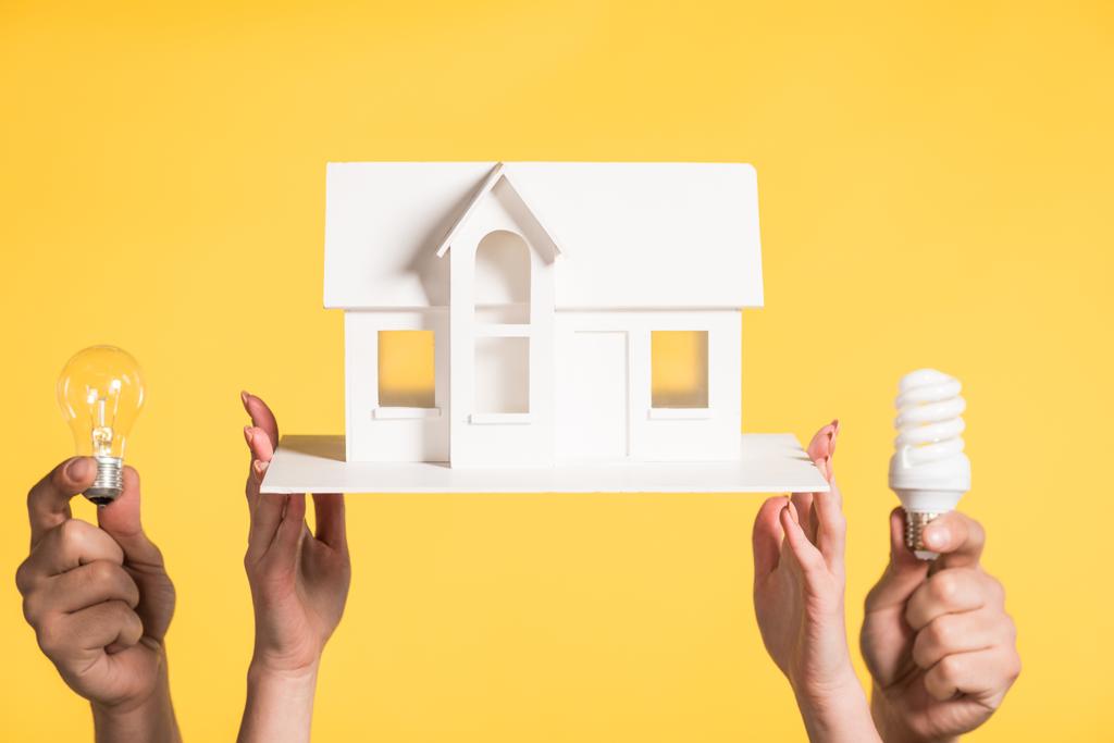 vista recortada de la mujer que sostiene el modelo de casa de cartón cerca del hombre con lámparas led y fluorescentes aisladas en amarillo, eficiencia energética en el hogar concepto
 - Foto, imagen