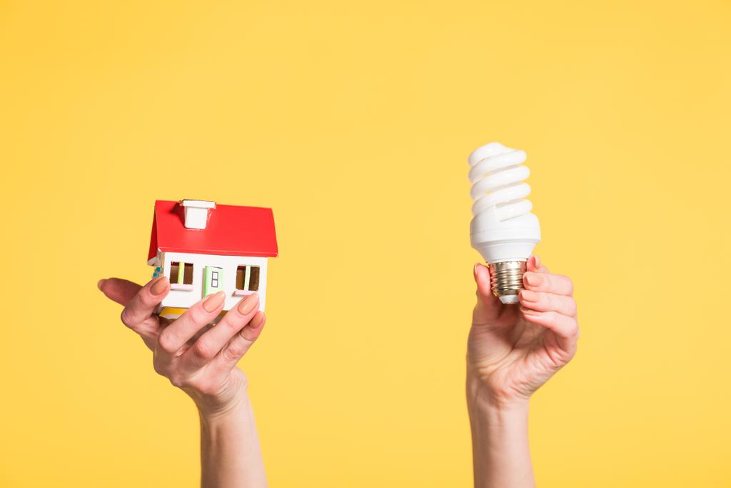 vista cortada da mulher segurando lâmpada fluorescente e modelo de casa em mãos isoladas em amarelo, eficiência energética no conceito de casa
 - Foto, Imagem