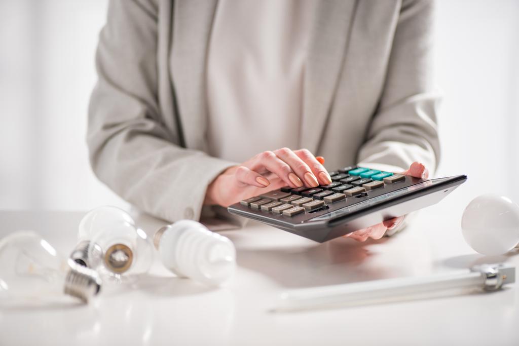 cropped view of woman using calculator near lamps on white background, energy efficiency concept - Photo, Image