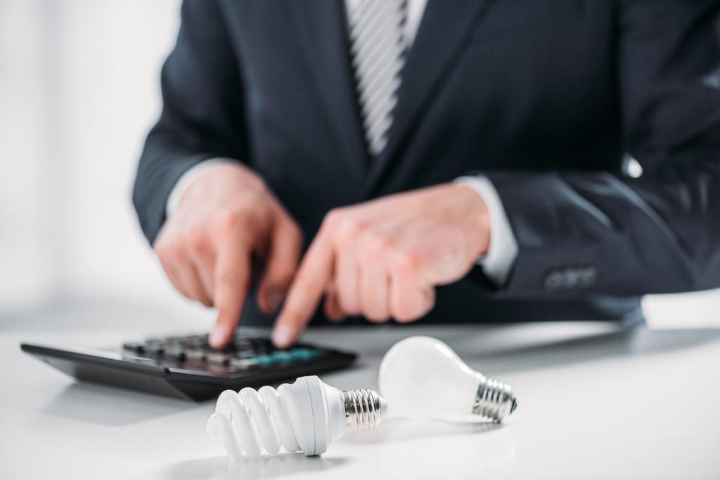 enfoque selectivo de lámparas fluorescentes cerca de hombre de negocios en traje usando calculadora sobre fondo blanco, concepto de eficiencia energética
 - Foto, Imagen