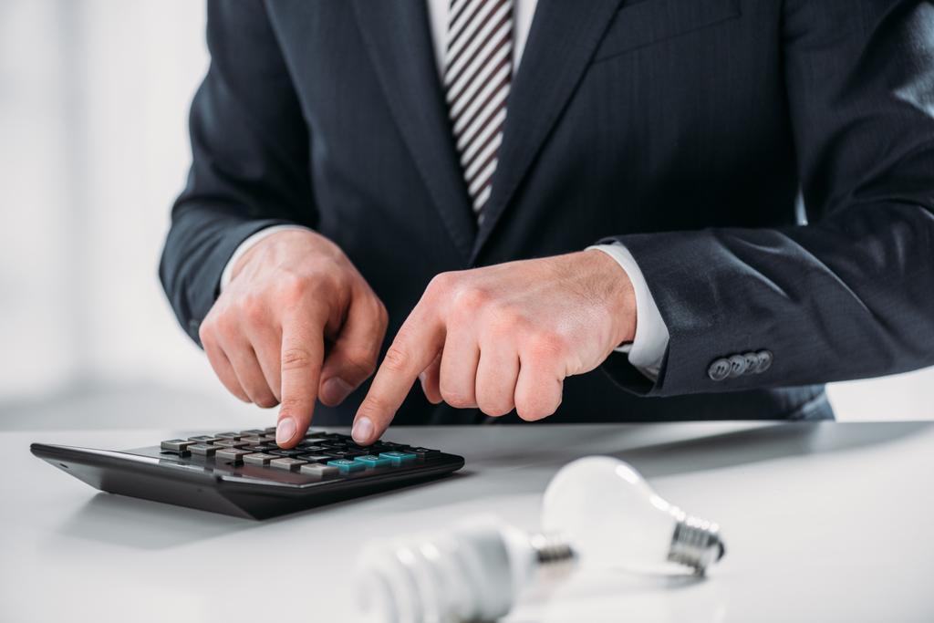 vista cortada do homem de negócios no terno usando calculadora perto de lâmpadas fluorescentes no fundo branco, conceito de eficiência energética
 - Foto, Imagem