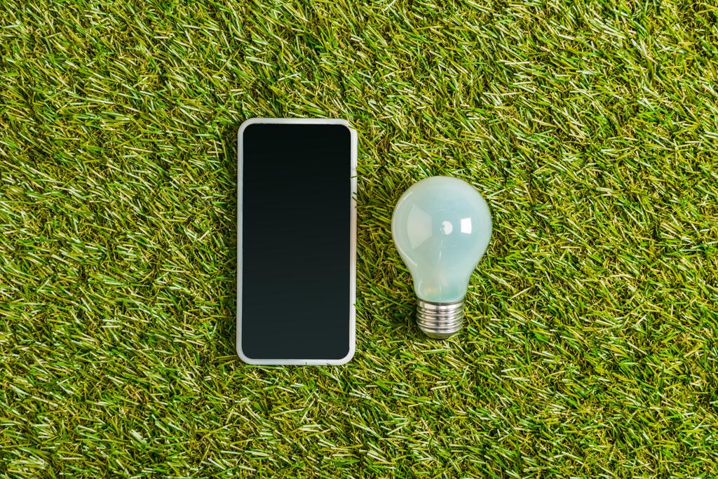 top view of fluorescent lamp near smartphone with blank screen on green grass, energy efficiency concept  - Photo, Image