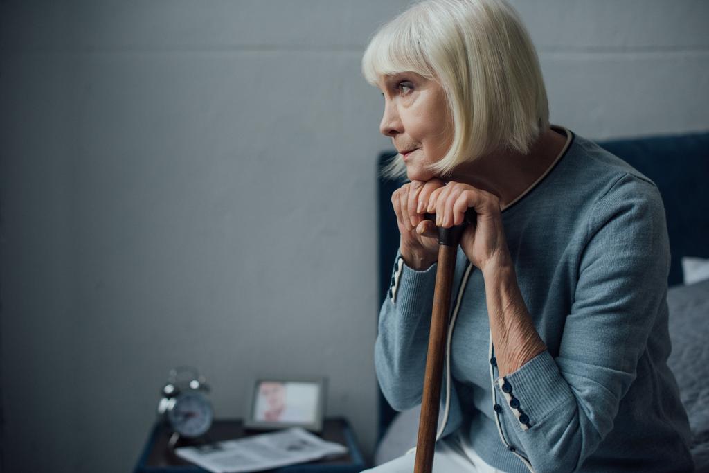 smutný starší žena s rukama na vycházkovou hůl, sedí na posteli doma a hledat dál - Fotografie, Obrázek