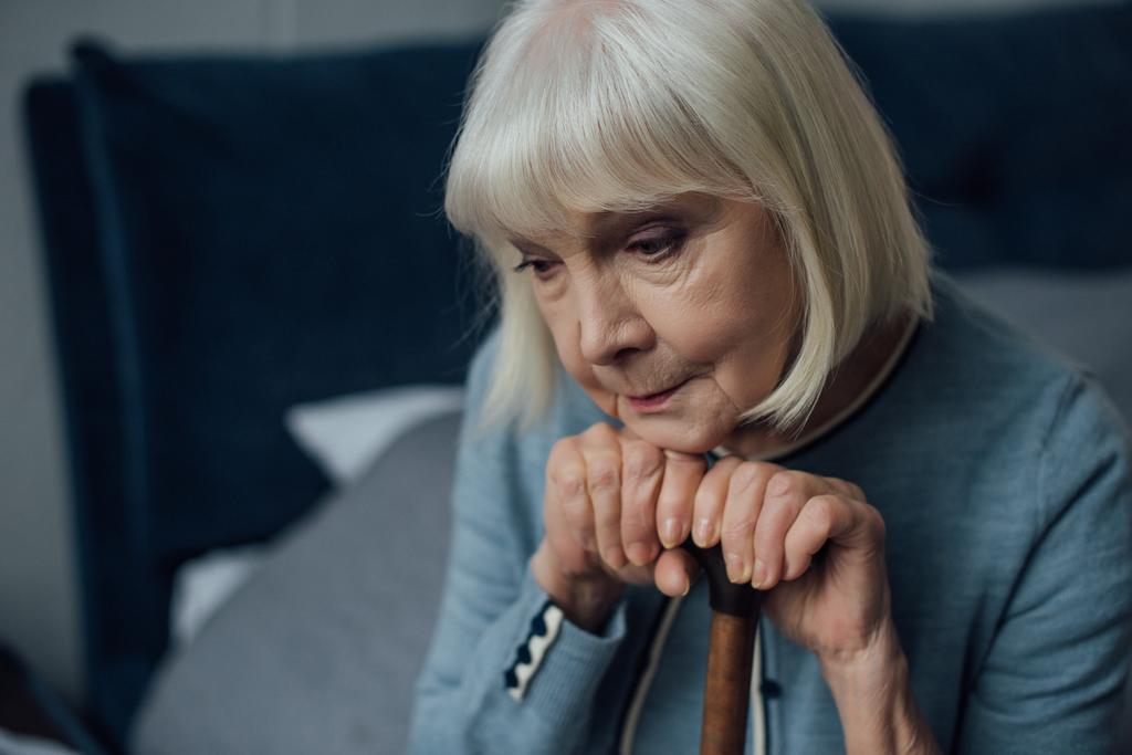portrait of upset senior woman with hands on walking stick sitting on bed at home - Photo, Image