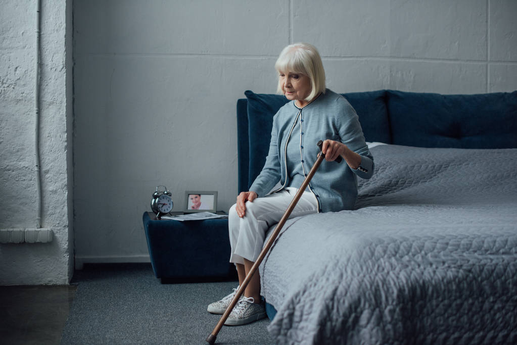 samotny zamyślony starszy kobieta siedzi na łóżku z laską w domu - Zdjęcie, obraz