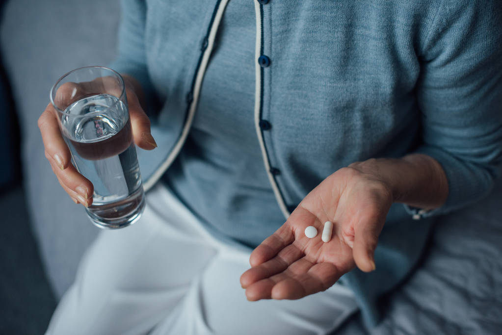 обрезанный вид пожилой женщины, сидящей и держащей таблетки со стаканом воды дома
 - Фото, изображение