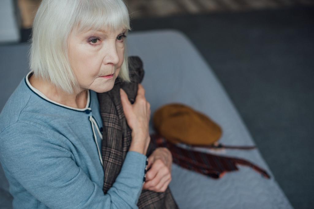 сумна старша жінка сидить на ліжку, дивиться вбік і тримає піджак вдома
 - Фото, зображення