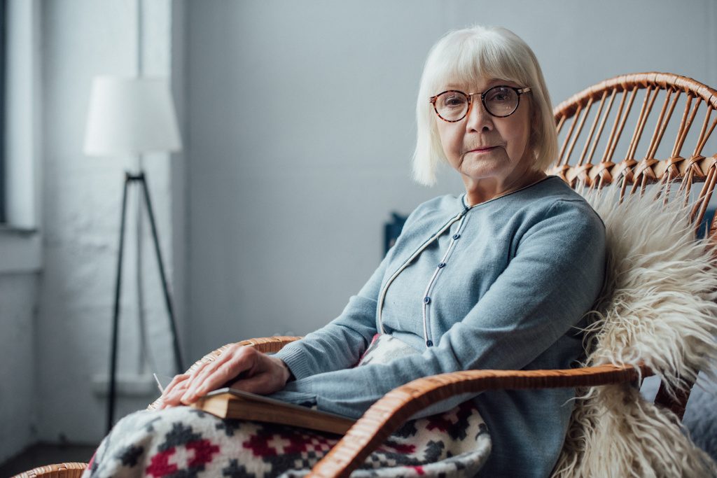 Starší žena sedí v proutěné houpací křeslo doma a při pohledu na fotoaparát - Fotografie, Obrázek