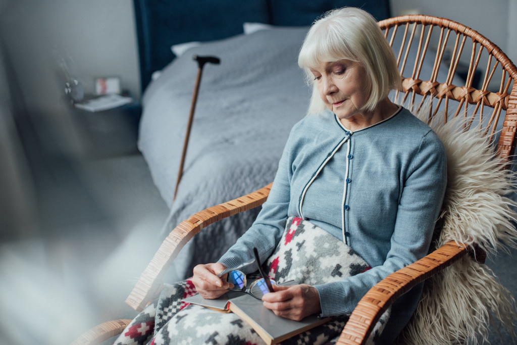 przemyślane starszy kobieta siedzi w wiklinowych bujane z książki w domu - Zdjęcie, obraz