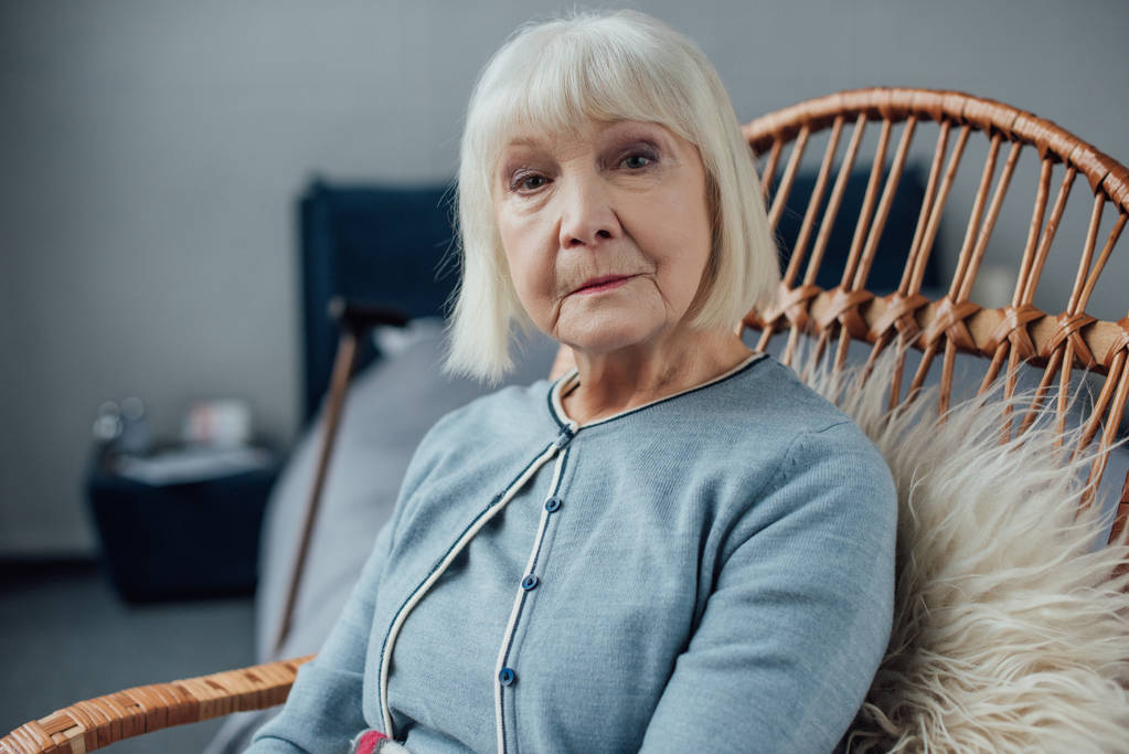 seriózní starší žena sedí v proutěné houpací křeslo doma a při pohledu na fotoaparát - Fotografie, Obrázek