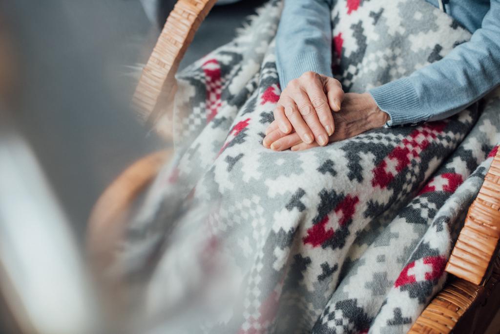 vista ritagliata della donna anziana seduta in vimini sedia a dondolo con coperta e mani piegate a casa
 - Foto, immagini