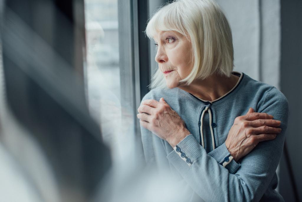 zamyšleně starší žena s překříženýma rukama při pohledu přes okno doma  - Fotografie, Obrázek