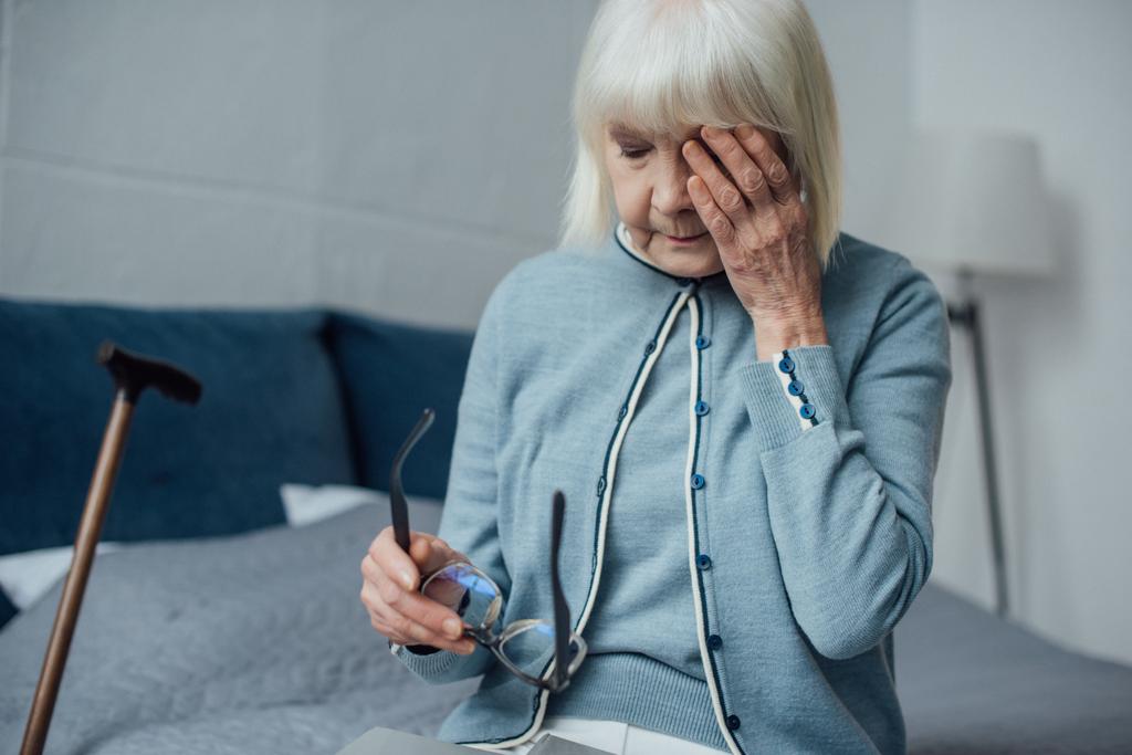 Traurige Seniorin mit Brille und grauem Haar wischt sich zu Hause Tränen und weint - Foto, Bild