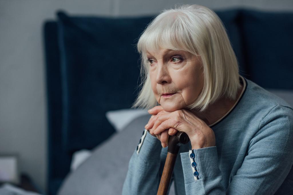 Nachdenkliche Seniorin mit den Händen am Gehstock sitzt zu Hause im Bett - Foto, Bild