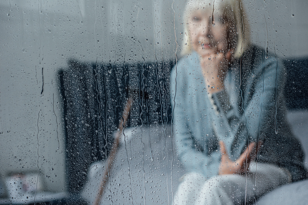 samotny senior kobieta siedzi na łóżku i wspierają podbródek ręką w domu przez okno z deszczu - Zdjęcie, obraz
