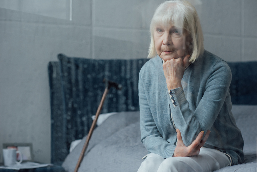 Traurige Seniorin sitzt zu Hause auf Bett und stützt Kinn mit der Hand - Foto, Bild