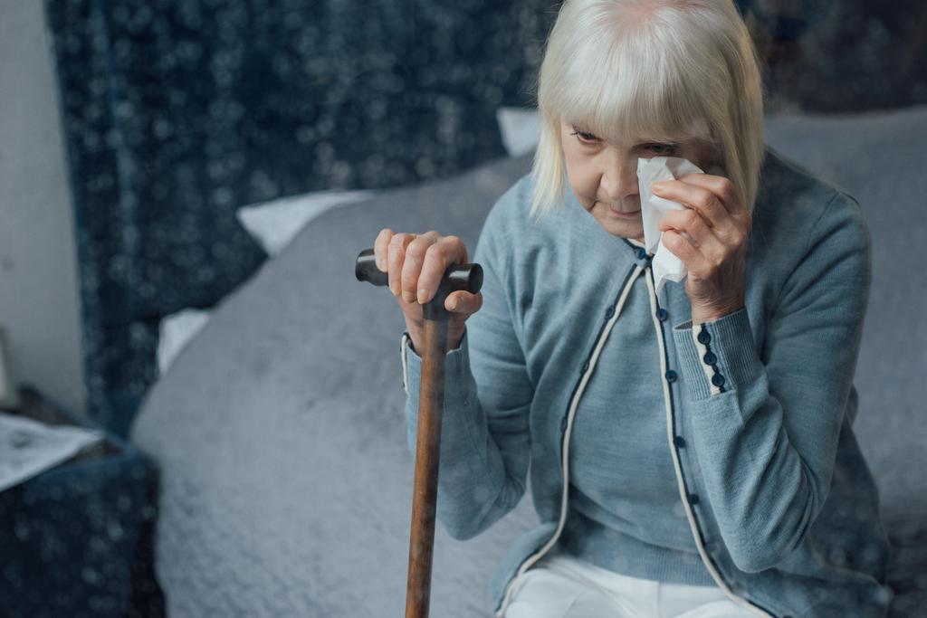 Ανώτερος γυναίκα κάθεται στο κρεβάτι, κρατώντας μπαστούνι και σκουπίζοντας τα δάκρυα με μαντήλι στο σπίτι - Φωτογραφία, εικόνα