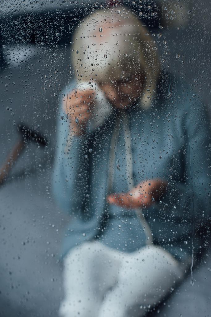 Ανώτερος γυναίκα κάθεται στο κρεβάτι και σκουπίζοντας τα δάκρυα με μαντήλι στο σπίτι μέσα από το παράθυρο με τις σταγόνες της βροχής - Φωτογραφία, εικόνα