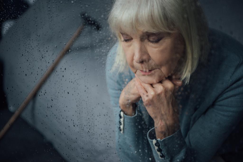 triste donna anziana seduta e appoggiando mento con le mani a casa attraverso la finestra con gocce di pioggia
 - Foto, immagini