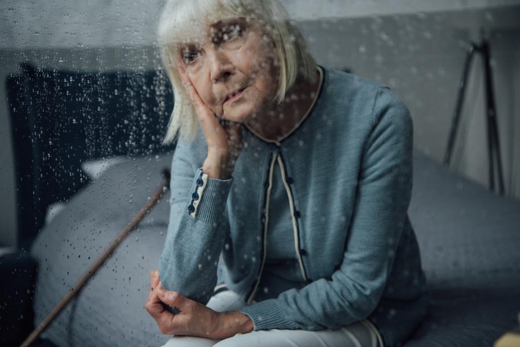 surullinen vanhempi nainen istuu sängyllä kotona ikkunan läpi sadepisaroilla
 - Valokuva, kuva