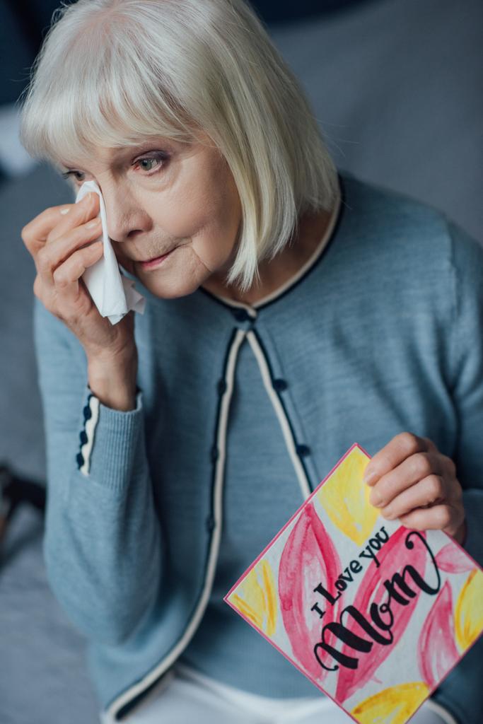 senior woman holding card with 'i love you mom' lettering, crying and wiping tears with handkerchief at home - Photo, Image