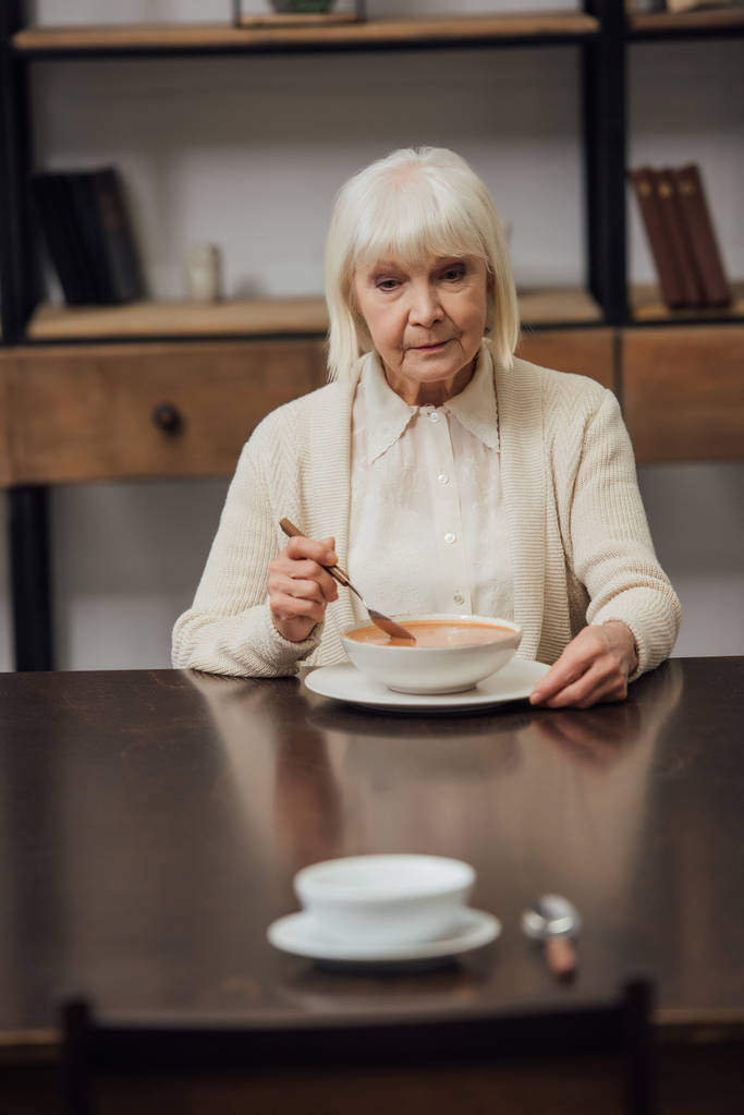 foco seletivo de mulher idosa triste comer à mesa com tigela e colher em primeiro plano
 - Foto, Imagem