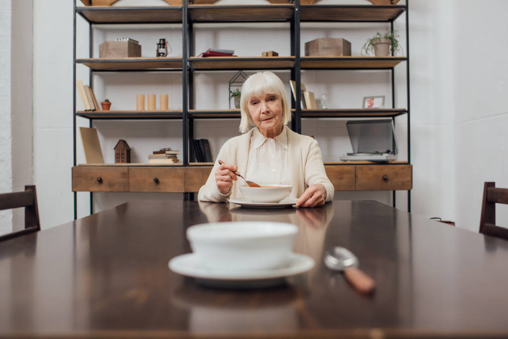 Selektivní fokus rozčilený starší ženy jíst u stolu s misku a lžíci na popředí - Fotografie, Obrázek