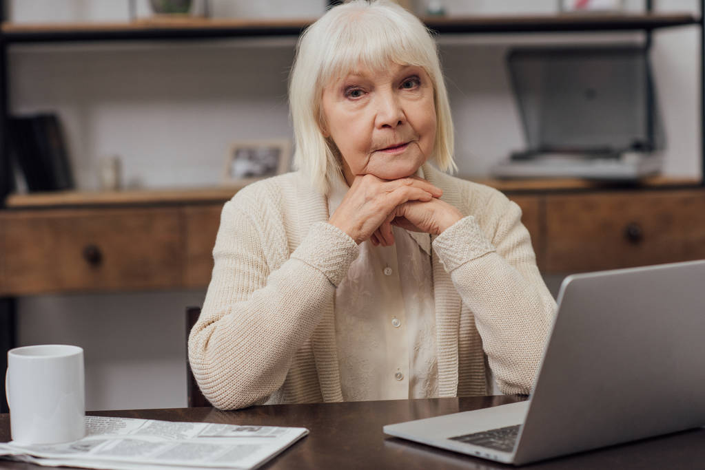 Seniorin sitzt mit Laptop am Tisch, blickt in Kamera und stützt Kinn mit den Händen ab - Foto, Bild