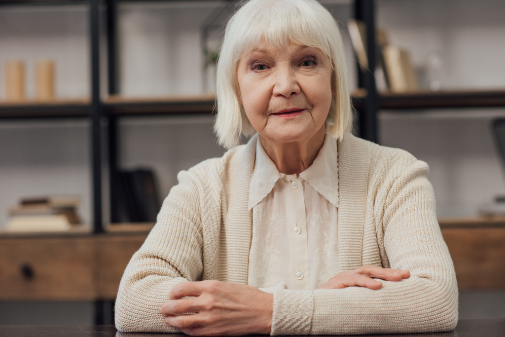 senior woman with grey hair and folded hands sitting looking at camera at home - Photo, Image