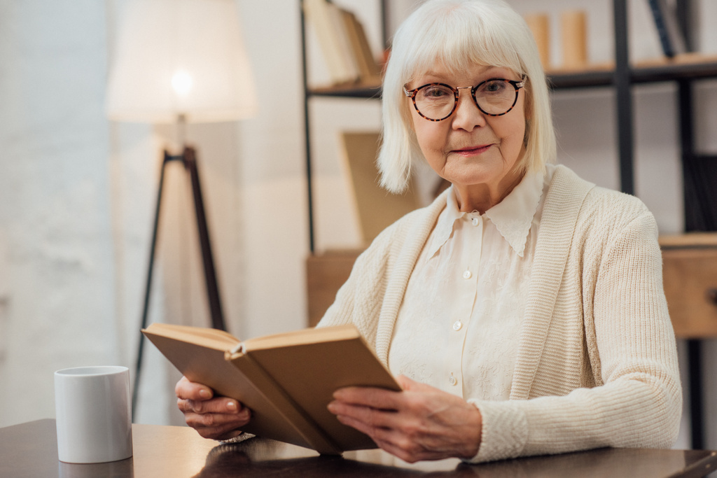 Senior kobietę w okularach siedzący przy stole i patrząc na kamery podczas czytania książki w domu - Zdjęcie, obraz