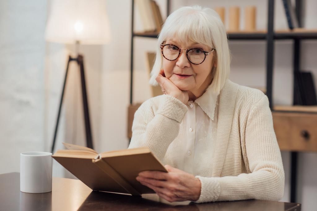 mulher sênior em óculos sentado à mesa, apoiando queixo com a mão e livro de leitura em casa
 - Foto, Imagem