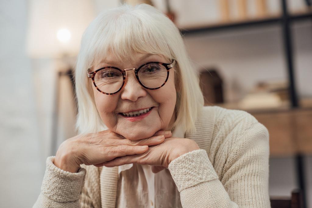 heureuse femme âgée avec cheveux gris appui menton avec les mains et en regardant la caméra à la maison
 - Photo, image
