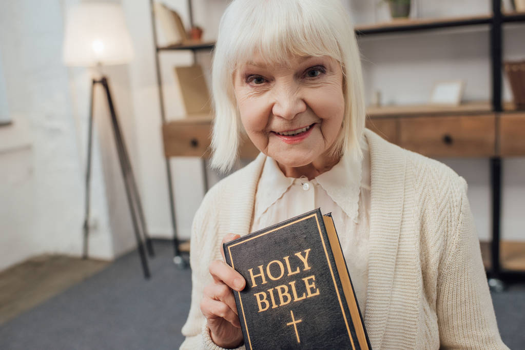 Lächelnde Seniorin blickt in die Kamera und hält die heilige Bibel zu Hause - Foto, Bild