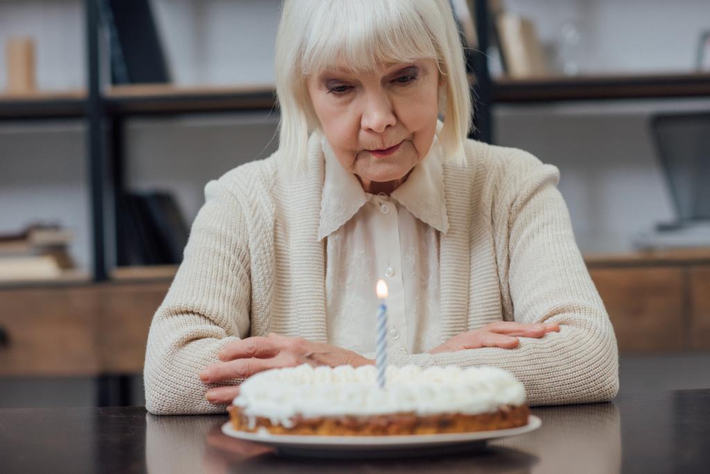 засмучена старша жінка сидить за столом і дивиться на торт на день народження з палаючою свічкою вдома
 - Фото, зображення