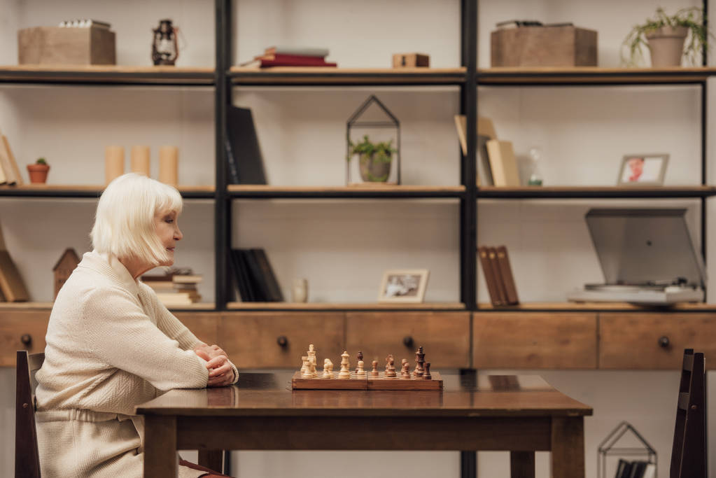 eenzame senior vrouw zitten aan tafel met schaakspel in woonkamer - Foto, afbeelding