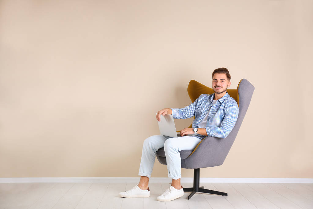 Красивий молодий чоловік з ноутбуком у кріслі біля кольорової стіни в приміщенні. Простір для тексту
 - Фото, зображення