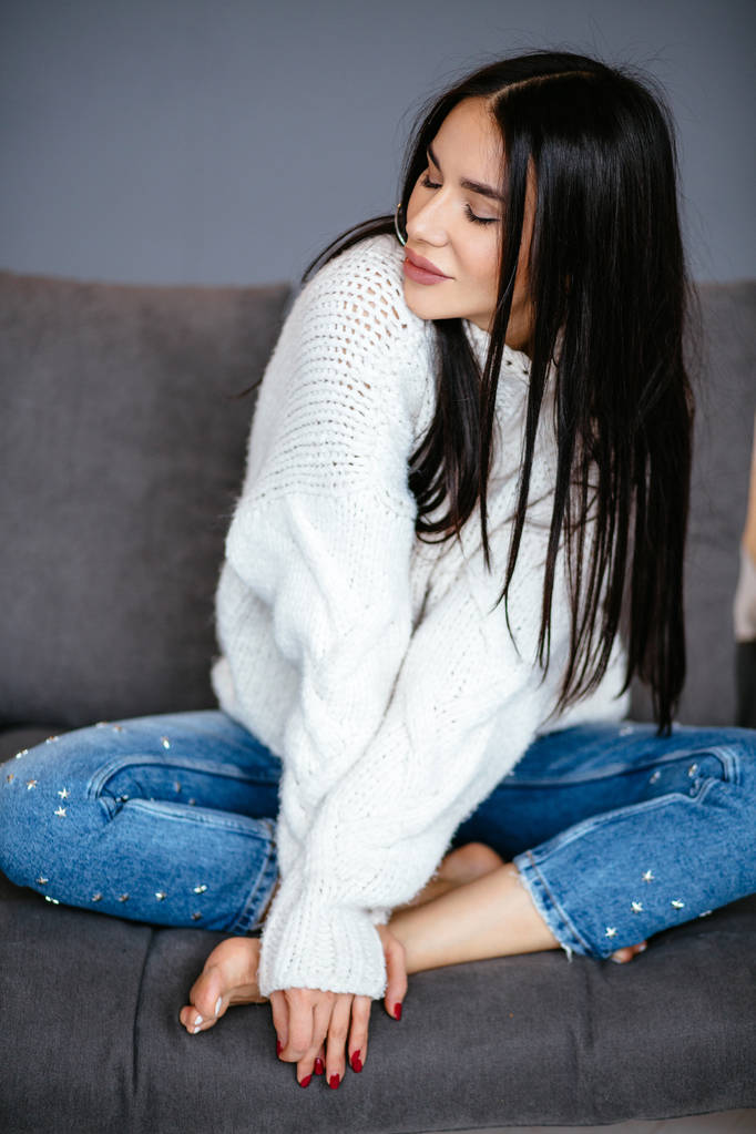 Hermosa chica en una chaqueta blanca y jeans está descansando en el sofá en casa. retrato, estilo escandinavo
 - Foto, imagen