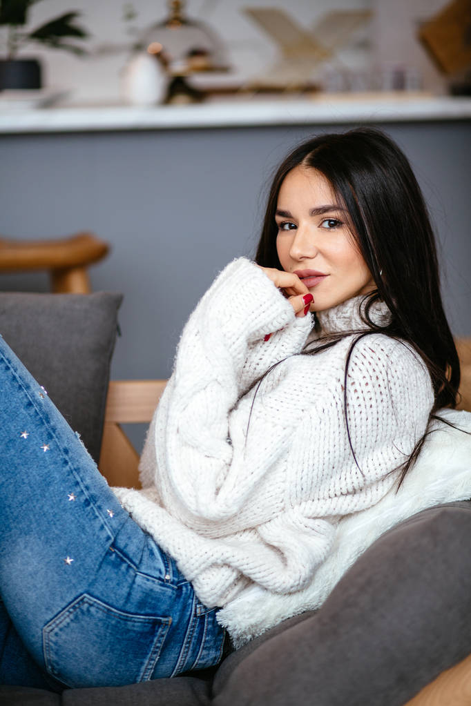 Красива дівчина в білій куртці і джинсах відпочиває на дивані вдома. портрет, скандинавський стиль
 - Фото, зображення