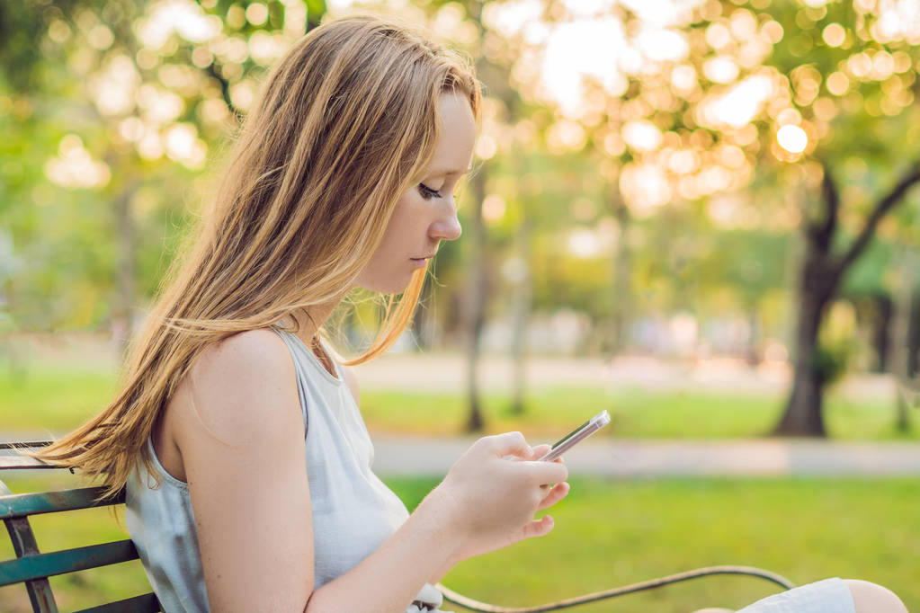 Joven chica tranquila usando su teléfono móvil para enviar un mensaje de texto mientras está de pie en un parque
 - Foto, imagen