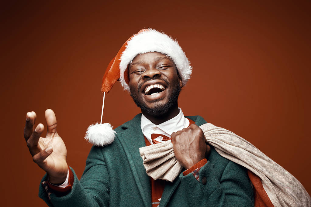 Papá Noel moderno. Sonriente hombre emocional posando en abrigo verde y suéter rojo, con sombrero y bolso de santa. Estudio plano, fondo marrón
 - Foto, Imagen