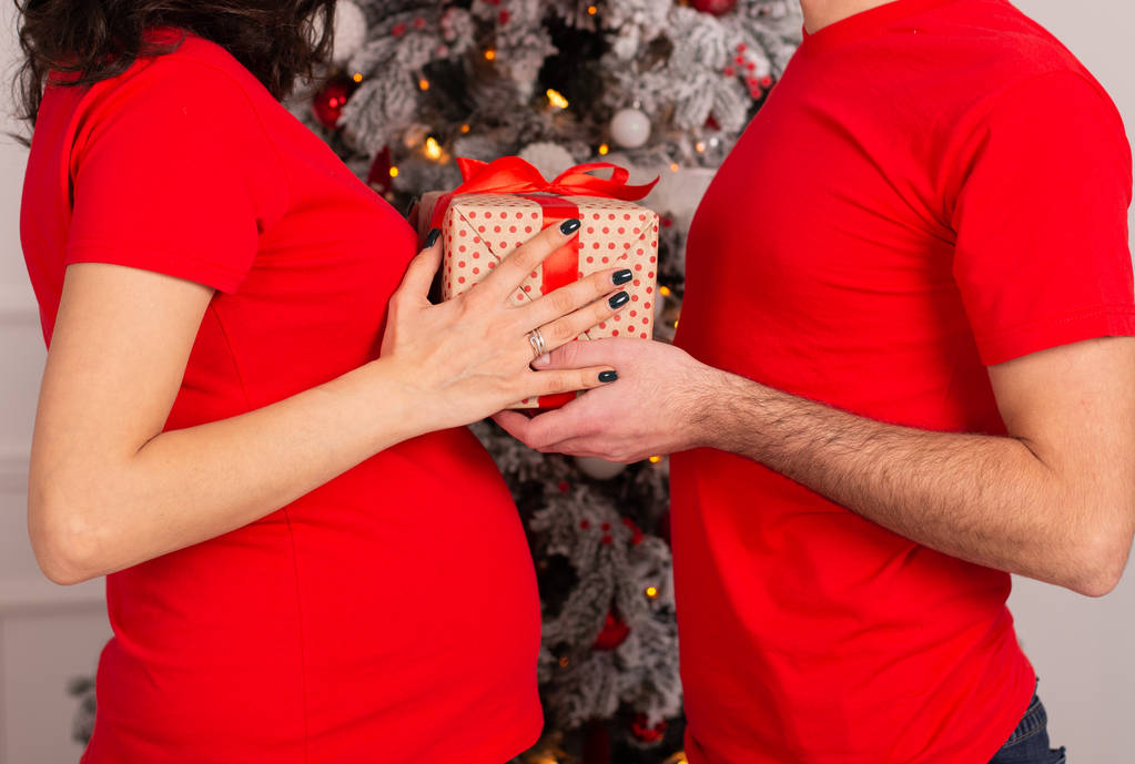 μερική του ανθρώπου δίνοντας παρόν στην έγκυο γυναίκα κοντά στο στολισμένο χριστουγεννιάτικο δέντρο  - Φωτογραφία, εικόνα