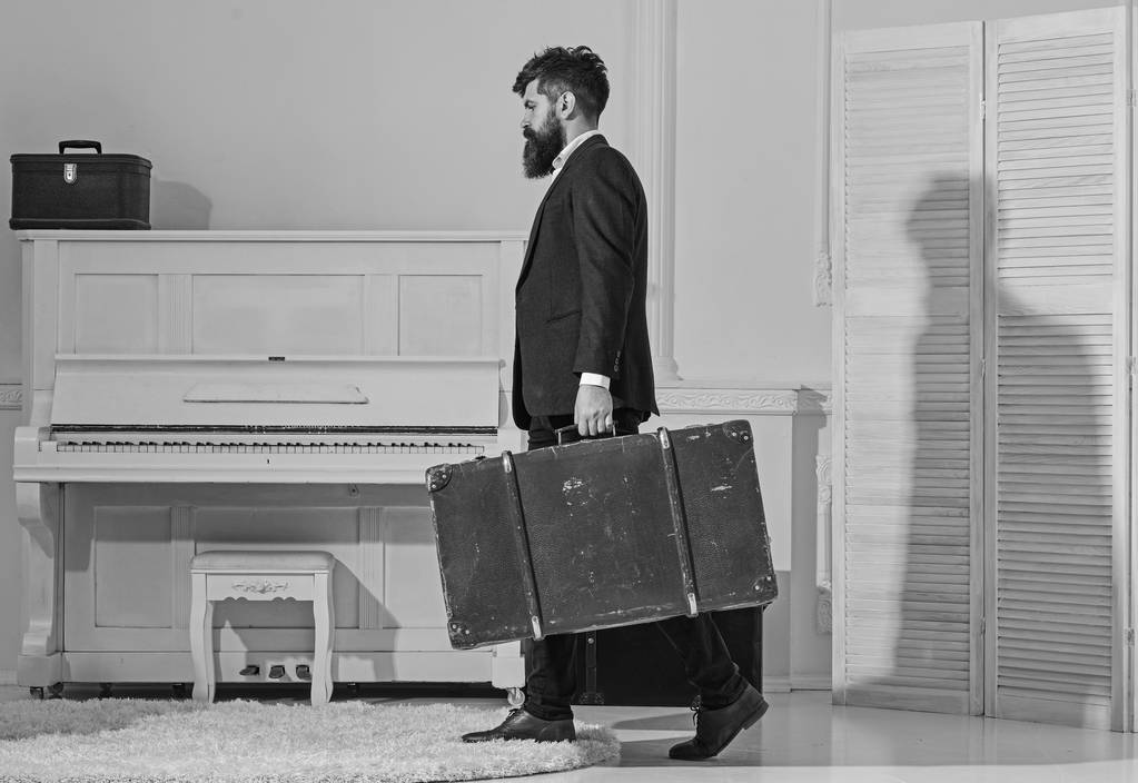 Macho stylish on strict face walks and carries big vintage suitcase, side view. Baggage and travelling concept. Man, traveller with beard and mustache with baggage, luxury white interior background - Photo, Image