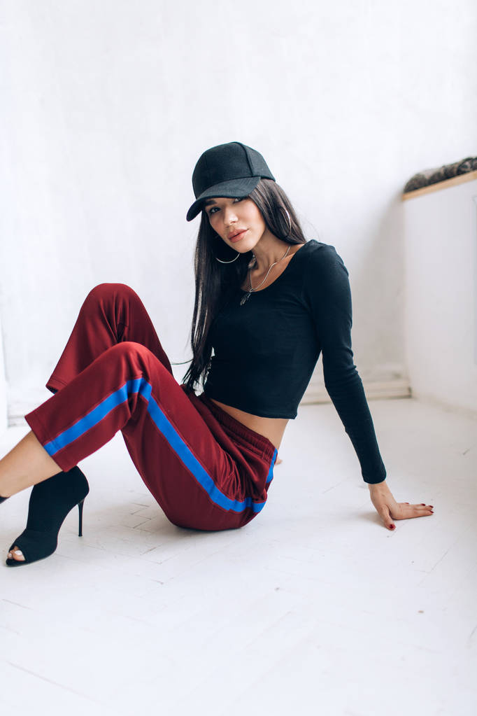 Portrait of a brunette girl in a cap and sportswear. Natural light. Red sweatpants, black top, black cap. - Photo, Image