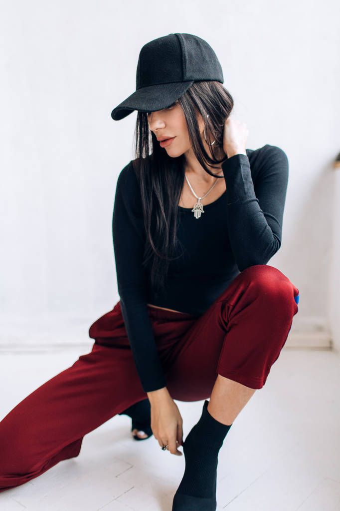 Portret van een brunette meisje in een cap en sportkleding. Natuurlijk licht. Rode trainingsbroek, zwart top, zwarte pet. - Foto, afbeelding